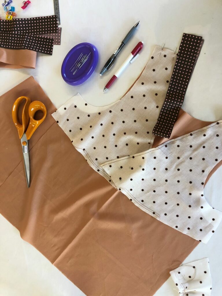 Sont posés sur la table de découpe de l'atelier, les ciseaux, épingles, crayons et les tissus qui composent le projet de nuisette féminin. Le tissu du haut est différent de celui sous la poitrine. Le biais d'un autre coloris également est prêt à être cousu sur l'encolure.