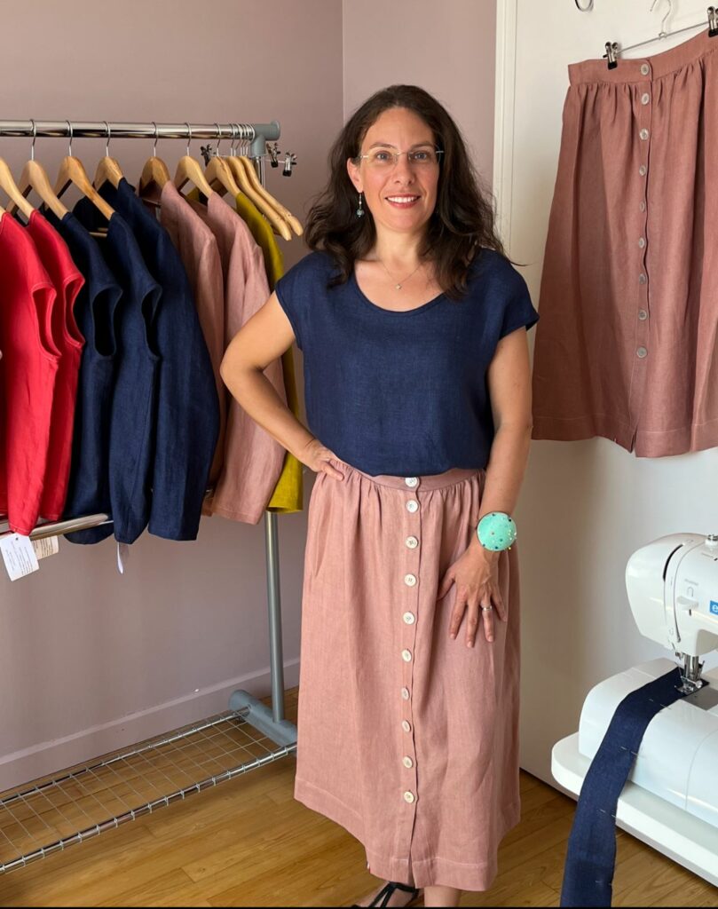 La créatrice Sophie Briand est dans son atelier debout à côté d'une machine à coudre. Derrière elle un portant sur lequel sont exposés fièrement ses créations de vêtements en lin pour femme (t-shirts, vestes et jupes longues). Elle porte elle-même une tenue fait-main en lin français : un t-shirt bleu marine et une jupe longue d'un coloris vieux rose.