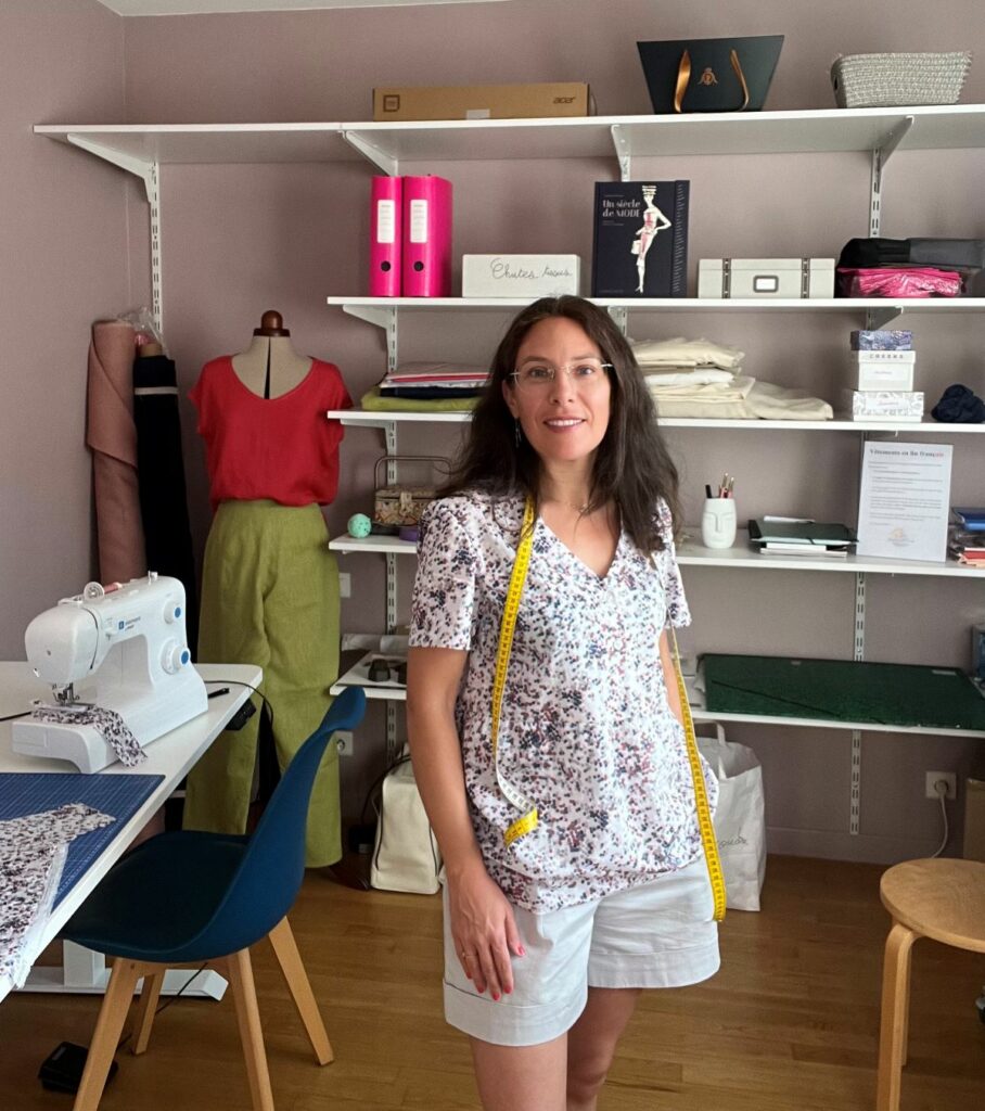 Sophie Briand décontractée debout dans son atelier de couture avec un mètre autour du cou. Derrière elle on aperçoit un mannequin portant un ensemble en lin, une machine à coudre et des étagères avec des tissus et des boîtes d'accessoires.