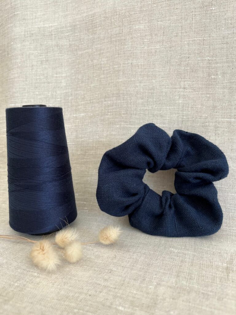 Un chouchou en lin bleu marine posé sur du lin beige avec une grosse bobine de fil assorti et des fleurs naturelles séchées à côté.