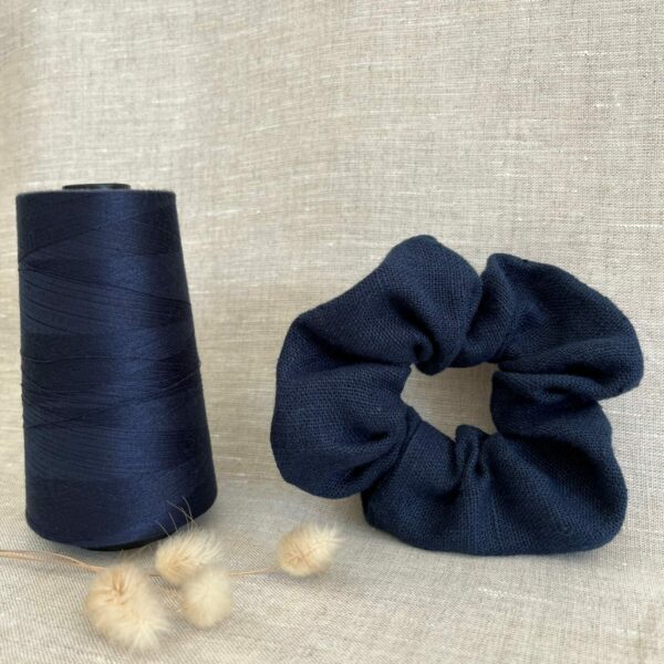 Un chouchou en lin bleu marine posé sur du lin beige avec une grosse bobine de fil assorti et des fleurs naturelles séchées à côté.
